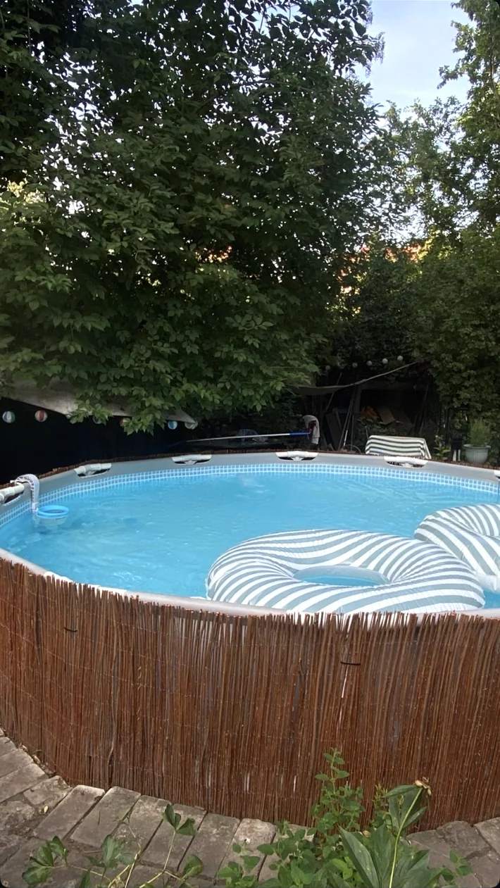 Round private pool with striped inflatable loungers, green trees and a wooden surround